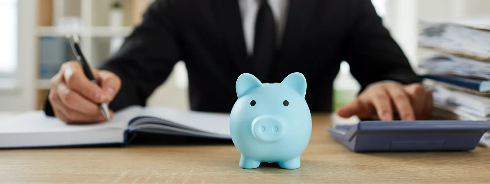 Man calculating a financial report and writing it, with a small blue piggy bank toy beside him, symbolizing savings and financial planning related to debt-to-income ratio.